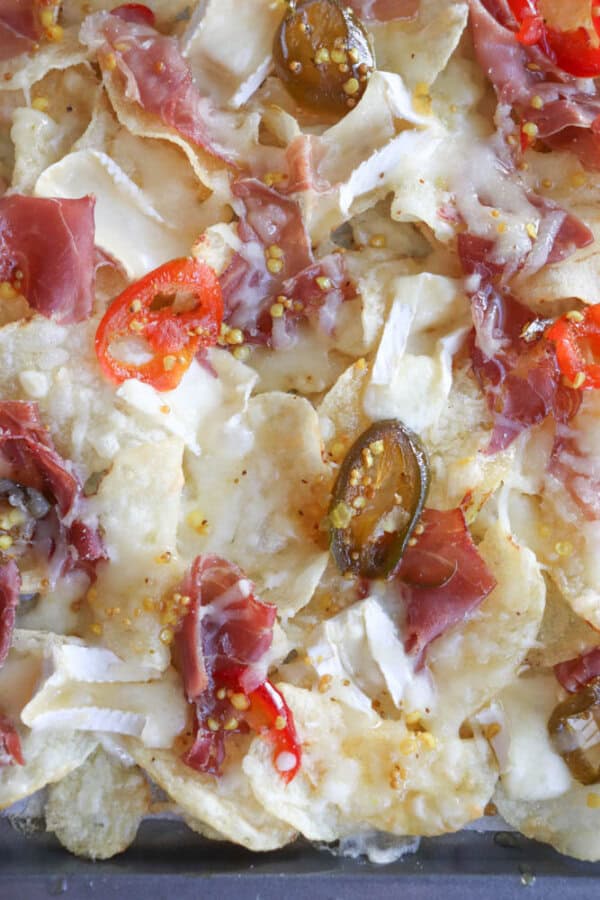 A close-up of loaded nachos featuring potato chips, melted cheese, sliced red and green peppers, and thinly sliced cured meat. The ingredients are spread over the chips, creating a colorful and appetizing dish.