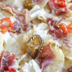 Close-up of nachos topped with melted cheese, sliced jalapeños, red peppers, and thin slices of cured meat. The ingredients are spread evenly, highlighting a variety of colors and textures on the tortilla chips.