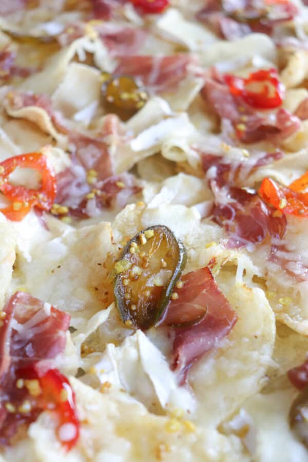 Close-up of nachos topped with melted cheese, sliced jalapeños, red peppers, and thin slices of cured meat. The ingredients are spread evenly, highlighting a variety of colors and textures on the tortilla chips.