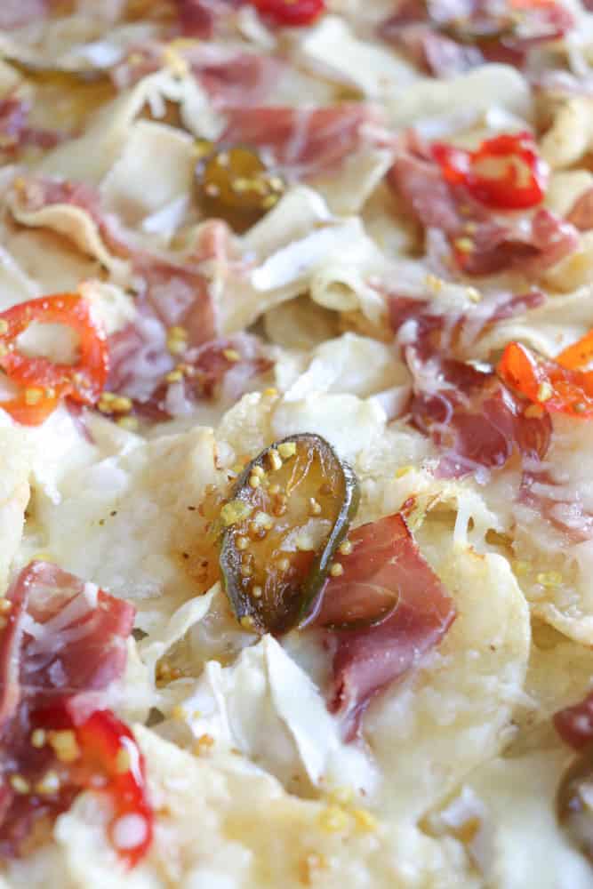 Close-up of nachos topped with melted cheese, sliced jalapeños, red peppers, and thin slices of cured meat. The ingredients are spread evenly, highlighting a variety of colors and textures on the tortilla chips.