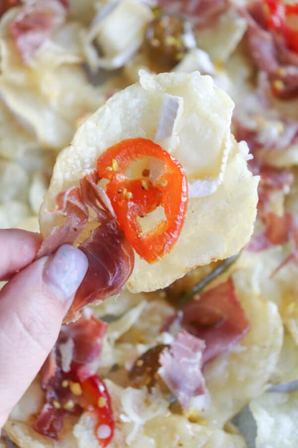 A hand holding a potato chip topped with a slice of red pepper, melted cheese, and prosciutto. The background shows more similarly topped chips.