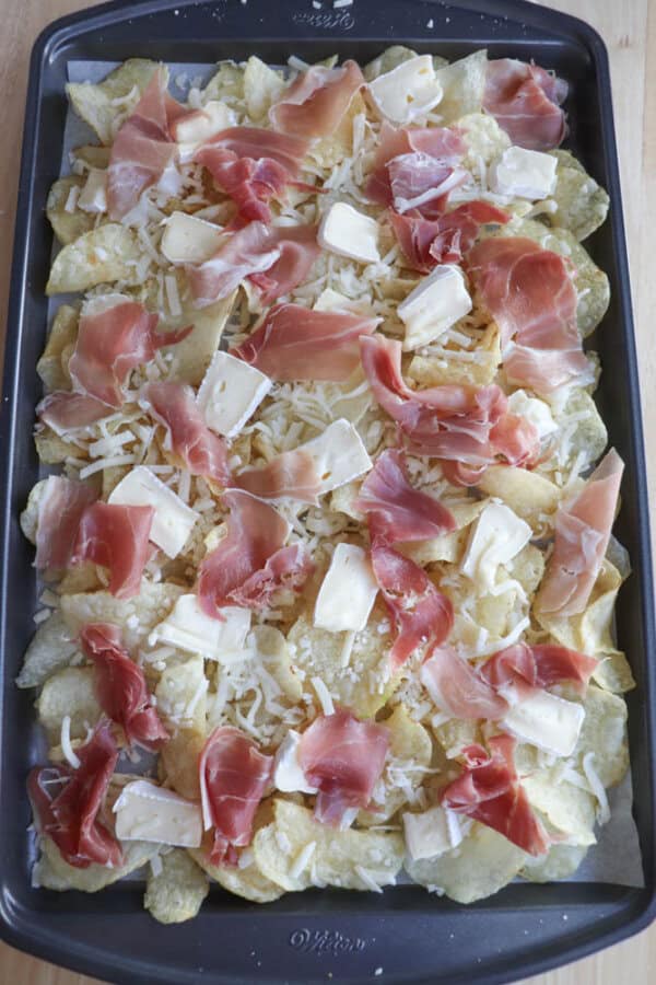 A baking tray filled with uncooked nachos. The tortilla chips are topped with slices of prosciutto, shredded cheese, and pieces of brie, arranged evenly across the surface.