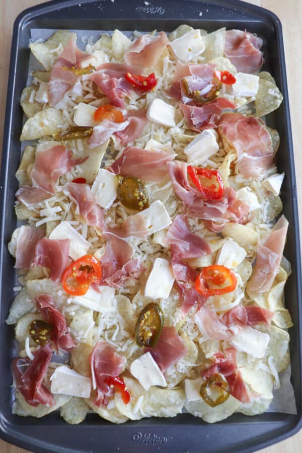 Sheet pan with tortilla chips topped with slices of prosciutto, jalapeño, halved cherry tomatoes, and scattered cheese.