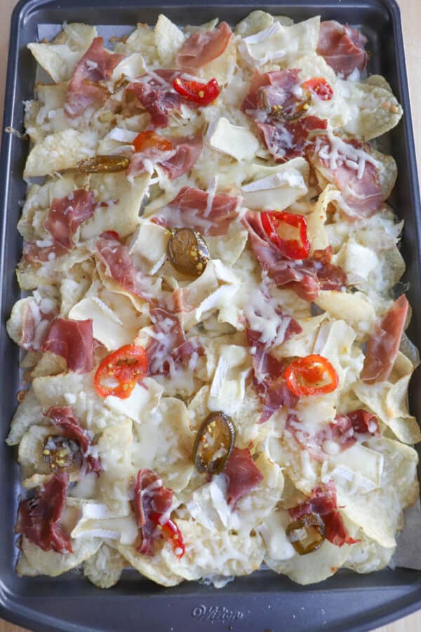 A tray of loaded nachos topped with melted cheese, slices of prosciutto, and sliced red chili peppers. The crispy chips are generously covered, creating a colorful and appetizing presentation.