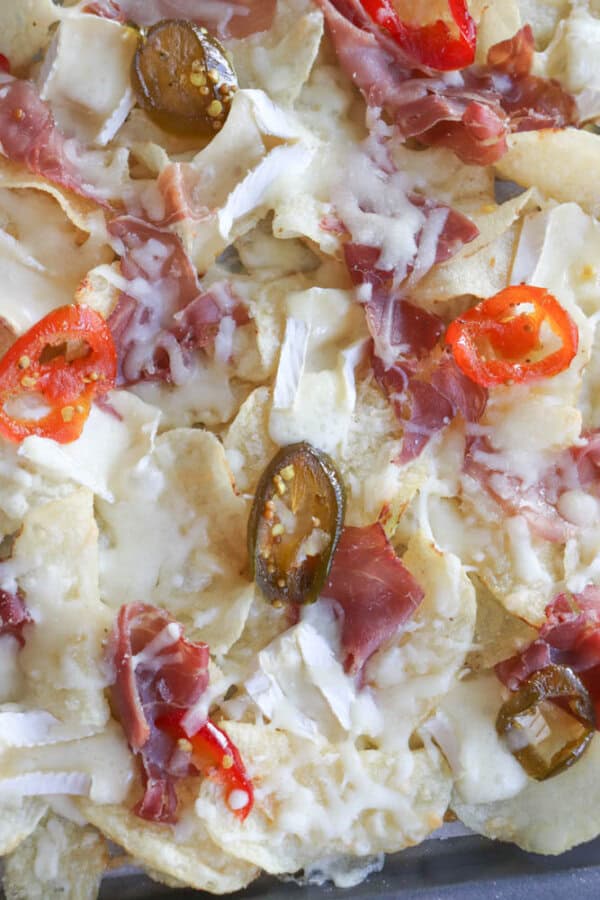 A close-up of loaded nachos topped with melted cheese, slices of ham, and jalapeño peppers. The dish features a mix of yellow corn tortilla chips and is garnished with thinly sliced red chili peppers.