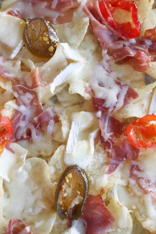 A close-up of nachos topped with prosciutto, melted cheese, jalapeño slices, and red bell pepper slices. The tortilla chips are layered with white cheese and thin slices of cured meat.