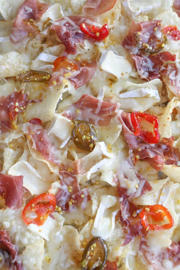 Close-up of nachos topped with melted cheese, thin slices of prosciutto, sliced red chili peppers, and jalapeños. The chips are slightly covered with crispy, melted cheese and bits of spicy toppings for a flavorful appearance.