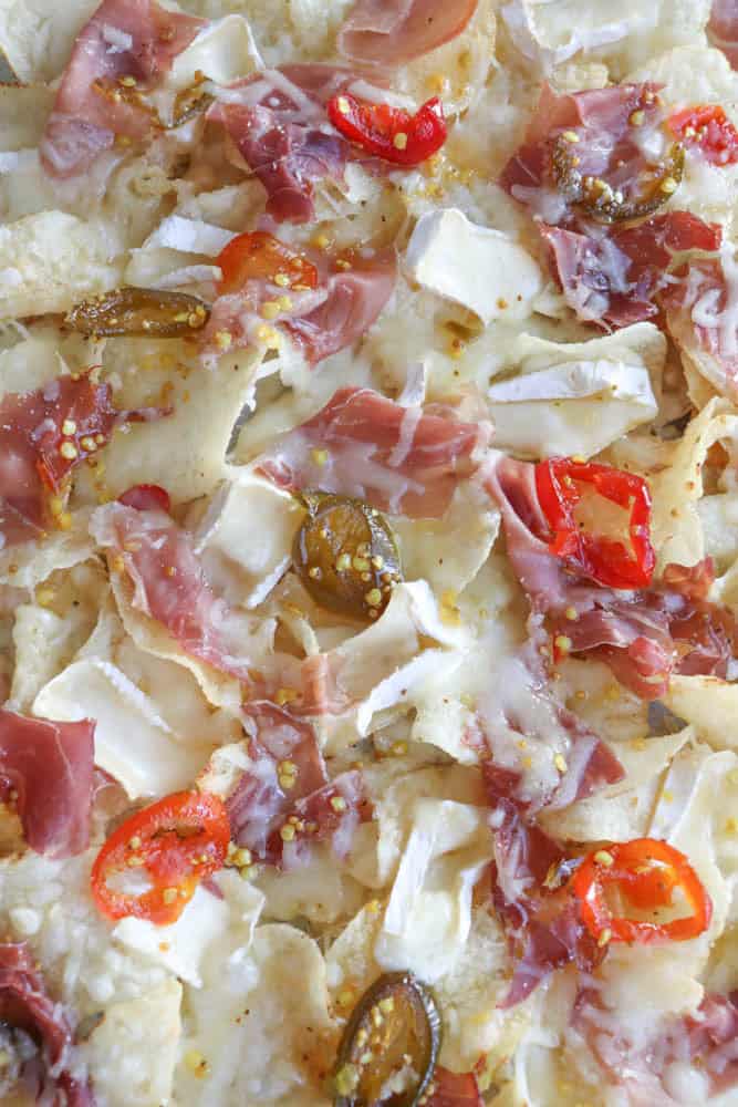 Close-up of nachos topped with melted cheese, thin slices of prosciutto, sliced red chili peppers, and jalapeños. The chips are slightly covered with crispy, melted cheese and bits of spicy toppings for a flavorful appearance.