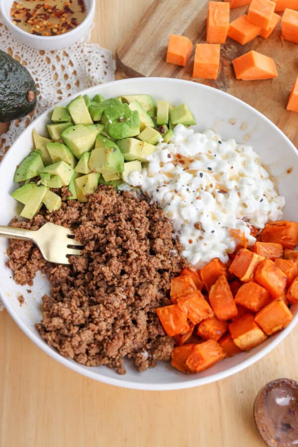 A white bowl filled with ground beef, diced avocado, cottage cheese, and roasted sweet potatoes. A fork is placed on the beef. Chopped sweet potatoes and half an avocado are on the wooden table beside it.