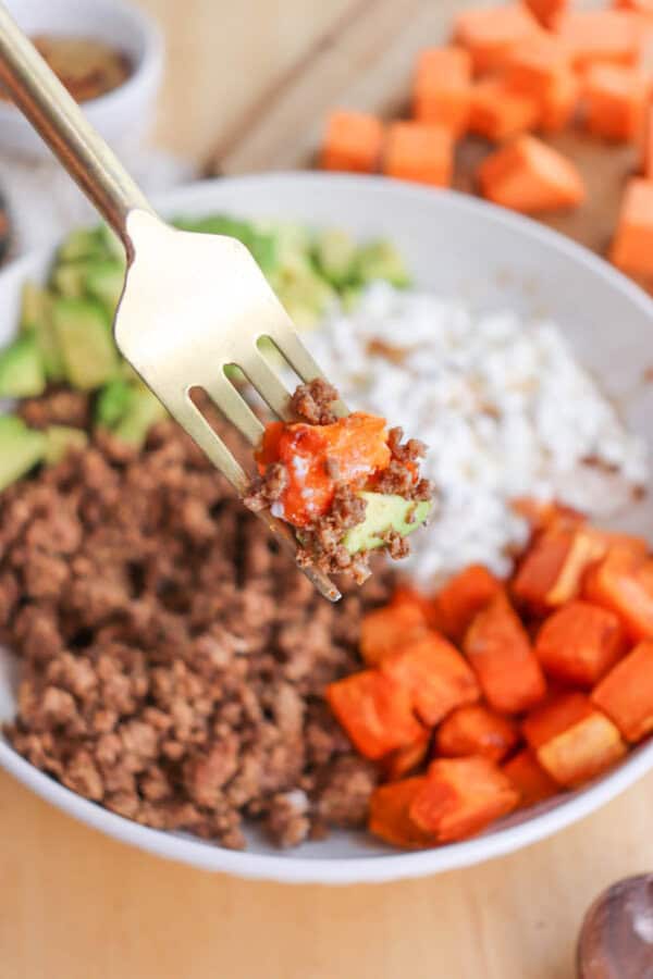 A fork holds a bite of sweet potato, avocado, and ground meat. In the background, a bowl contains sections of ground meat, diced sweet potatoes, avocado, and a white grain or cheese against a wooden surface.