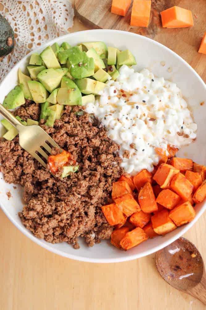 A bowl filled with diced avocado, cottage cheese, roasted sweet potato cubes, and cooked ground beef. A gold fork rests in the bowl, and theres a lace napkin partially visible underneath it. A wooden spoon and sweet potato cubes are nearby.