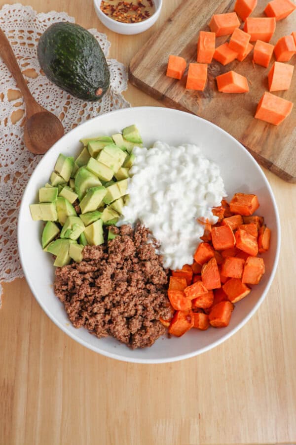 A bowl on a wooden table contains diced avocado, cottage cheese, cooked ground beef, and roasted sweet potatoes. An avocado and chopped sweet potatoes are nearby on a lace doily and a wooden cutting board. A small bowl of red pepper flakes is in the background.