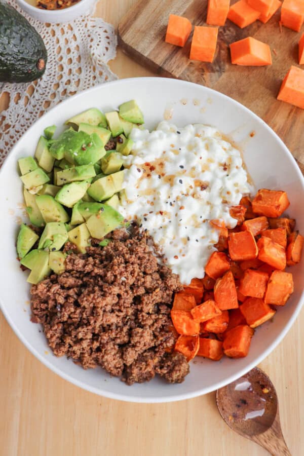 A bowl with chopped avocado, cottage cheese, cooked ground beef, and roasted sweet potatoes. The ingredients are arranged in sections, creating a colorful contrast. Additional sweet potato cubes and an avocado are visible in the background.