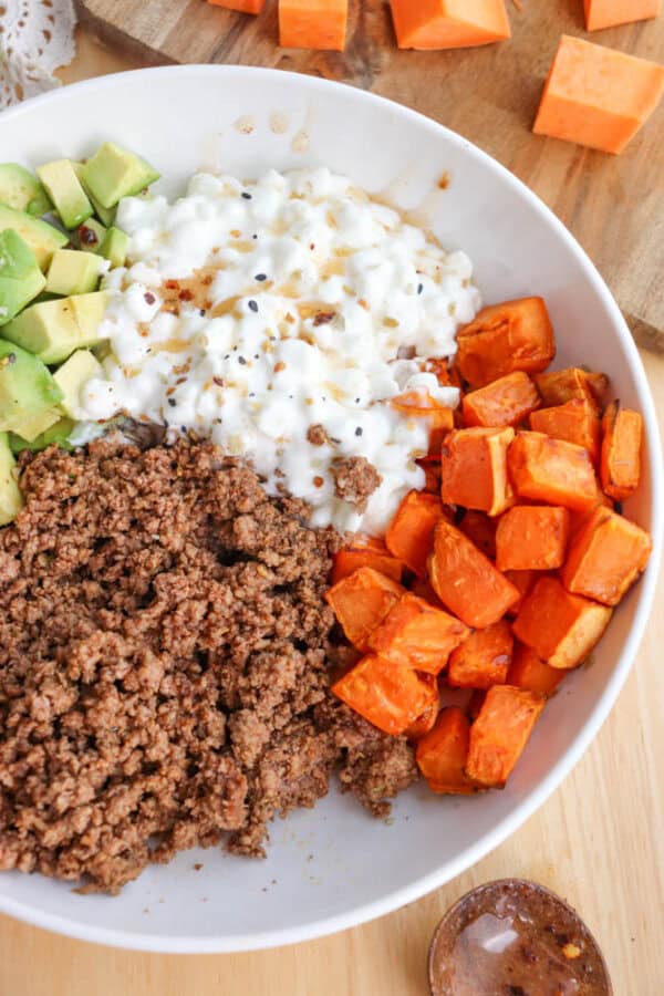 A bowl containing a mix of ingredients: ground meat, cubed sweet potatoes, diced avocado, and cottage cheese, sprinkled with seasoning. Additional cubed sweet potatoes are visible on a wooden board in the background.