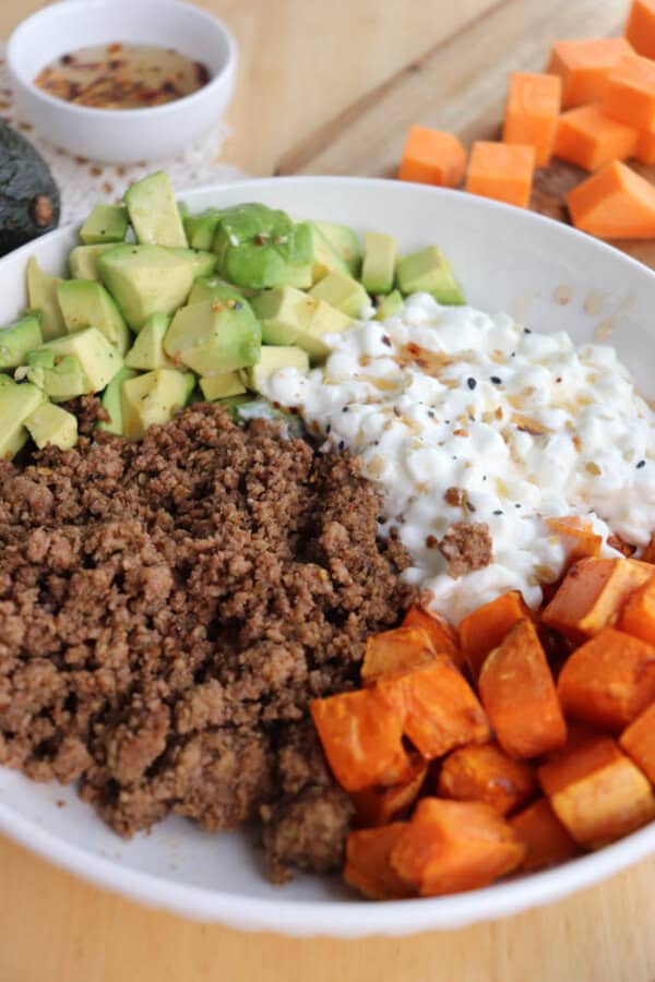 A bowl filled with ground beef, diced avocado, cottage cheese, and roasted sweet potato chunks. The background includes a small bowl of sauce and additional sweet potato pieces on a wooden surface.