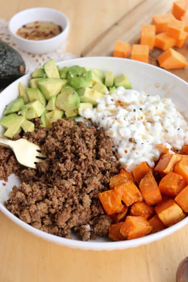 A bowl filled with cooked ground beef, diced avocado, cottage cheese, and roasted sweet potato cubes. A gold fork rests on the edge. A small bowl of seasoning and cubed sweet potatoes are on the table.