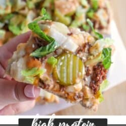 A hand holds a slice of Big Mac-inspired pizza topped with ground meat, pickles, onions, lettuce, cheese, and sesame seeds. More pizza slices are visible in the background. Text reads high protein BIG MAC PIZZA homemadeheather.com.