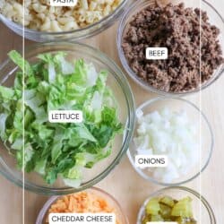 Bowls containing cooked pasta, sauce, ground beef, chopped lettuce, diced onions, shredded cheddar cheese, and sliced pickles are arranged on a wooden surface, each item labeled with text.