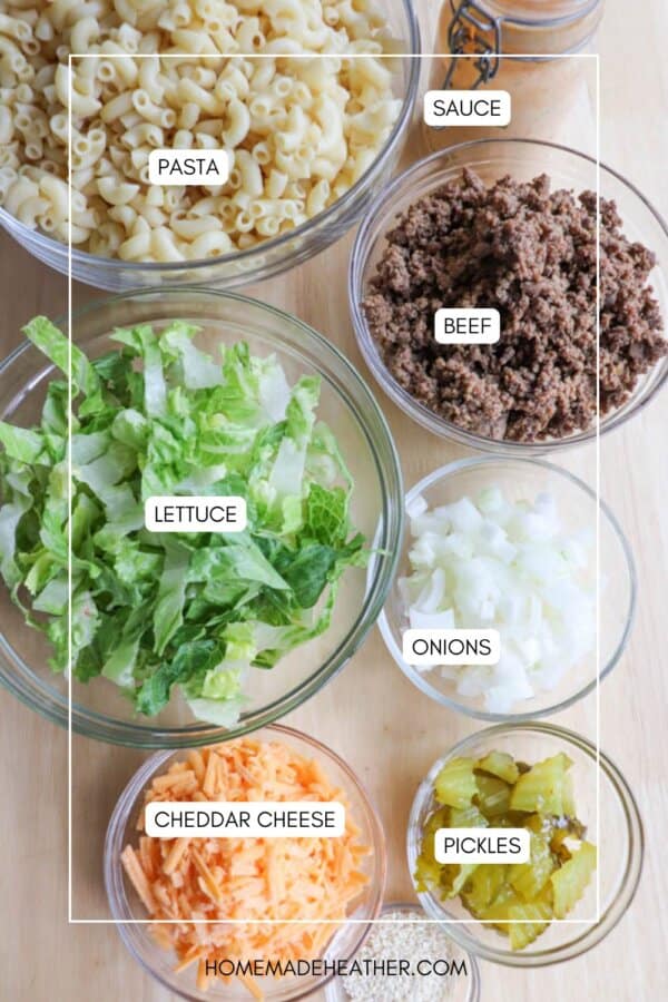 Bowls containing cooked pasta, sauce, ground beef, chopped lettuce, diced onions, shredded cheddar cheese, and sliced pickles are arranged on a wooden surface, each item labeled with text.