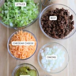 Six bowls on a wooden surface filled with ingredients for cheeseburger salad: chopped lettuce, cooked ground beef, shredded cheddar cheese, diced onions, sliced pickles, sesame seeds, and a jar of creamy sauce.