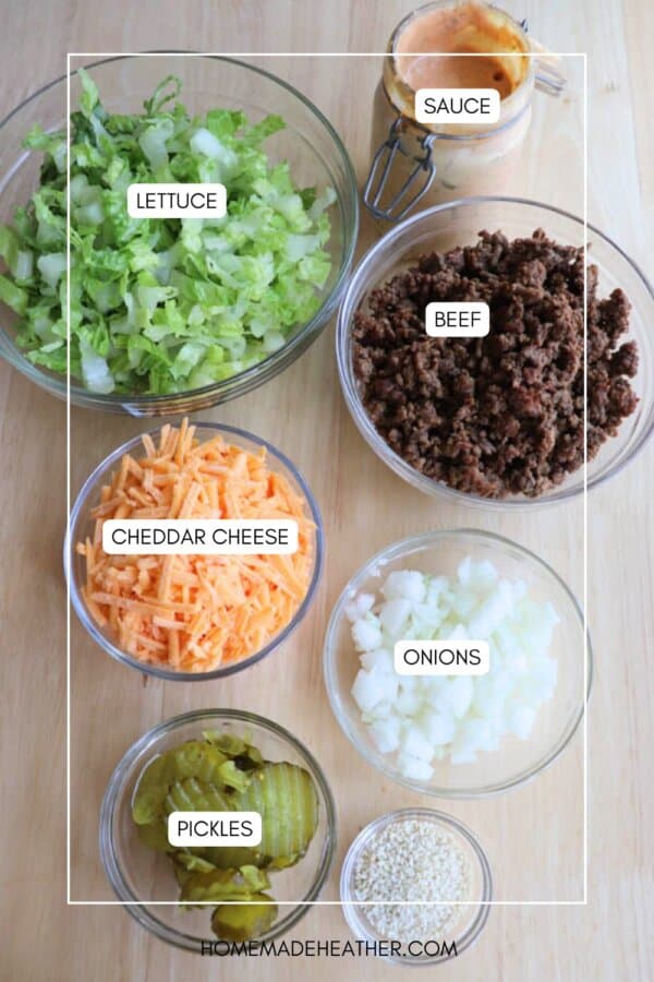 Six bowls on a wooden surface filled with ingredients for cheeseburger salad: chopped lettuce, cooked ground beef, shredded cheddar cheese, diced onions, sliced pickles, sesame seeds, and a jar of creamy sauce.