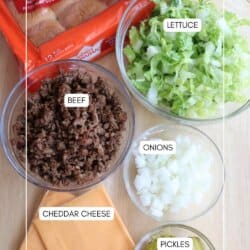 A top-down view of ingredients for sliders: a package of Hawaiian rolls, a bowl of shredded lettuce, cooked ground beef, diced onions, sliced cheddar cheese, and chopped pickles, all arranged on a table.