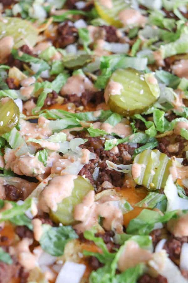 Close-up of a dish topped with ground beef, shredded lettuce, sliced pickles, diced onions, melted cheese, and a creamy sauce, resembling the toppings of a cheeseburger.