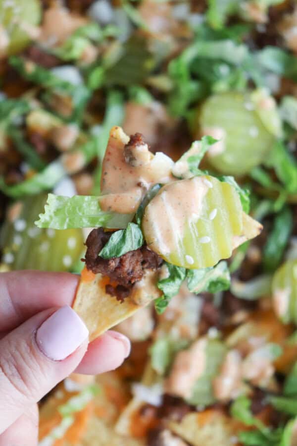 A hand holding a tortilla chip topped with ground beef, shredded lettuce, sliced pickles, and a drizzle of creamy Big Mac sauce, with more nachos blurred in the background.