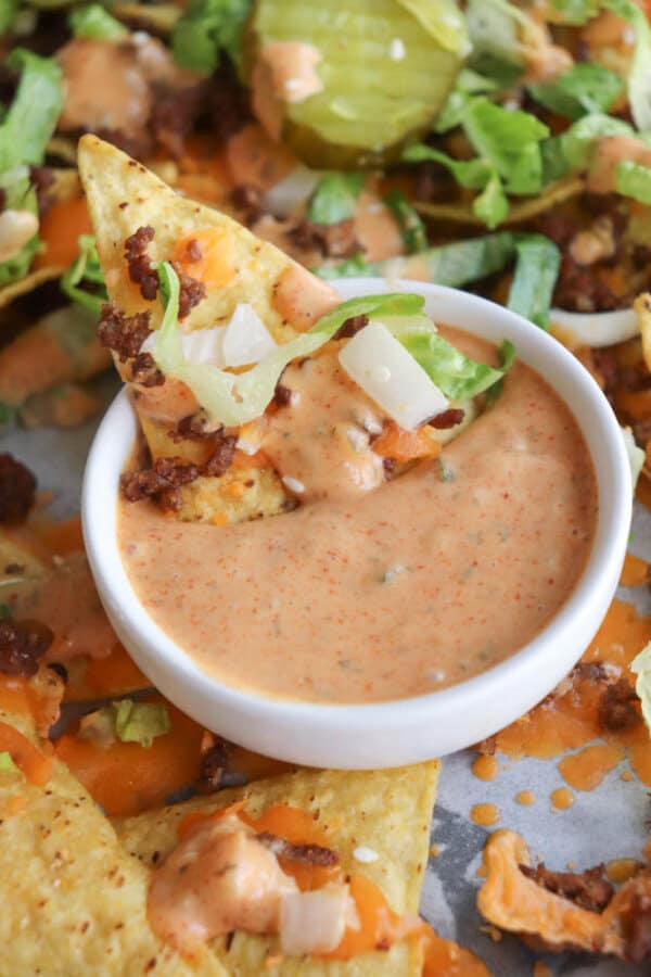 A tortilla chip topped with lettuce, onions, and ground beef is dipped in a small bowl of creamy, Big Mac sauce. More nachos with similar toppings are visible in the background.