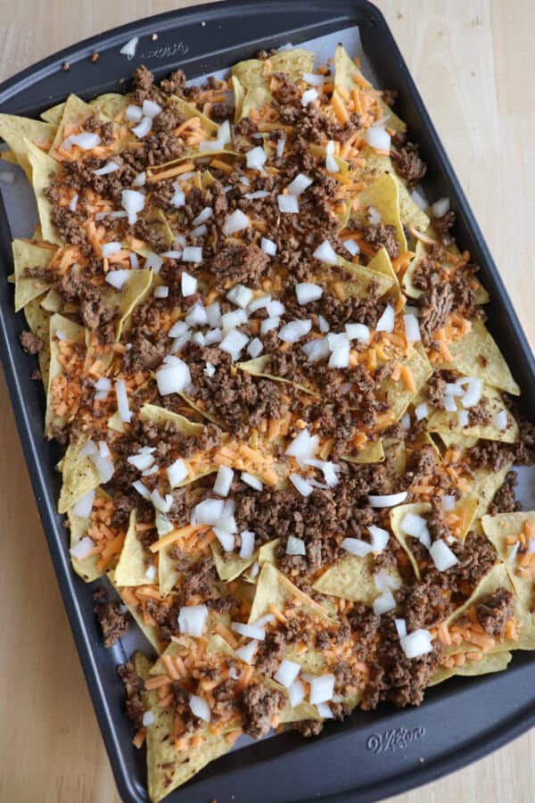 A baking tray filled with nachos topped with ground beef, shredded cheddar cheese, and chopped raw onions, ready to be baked.