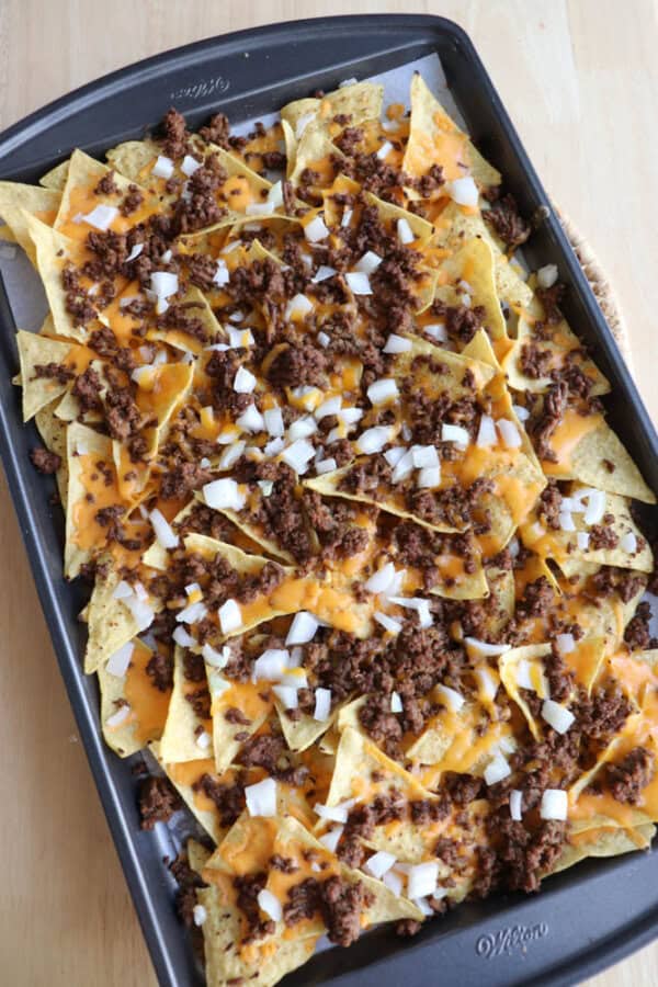A baking tray filled with tortilla chips topped with melted cheddar cheese, cooked ground beef, and chopped onions, ready to serve.