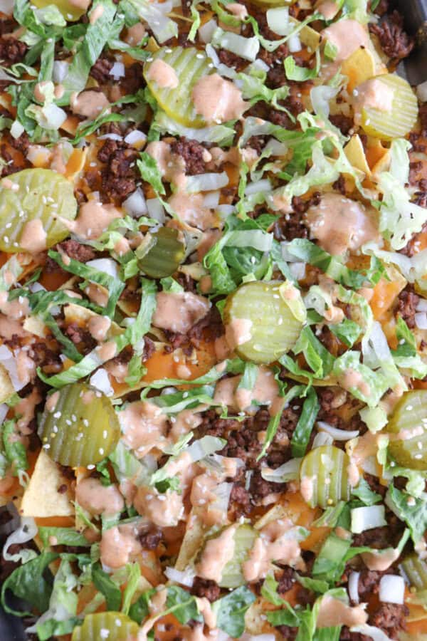 A close-up of loaded nachos topped with ground beef, shredded lettuce, sliced pickles, diced onions, melted cheese, and a drizzle of creamy sauce.