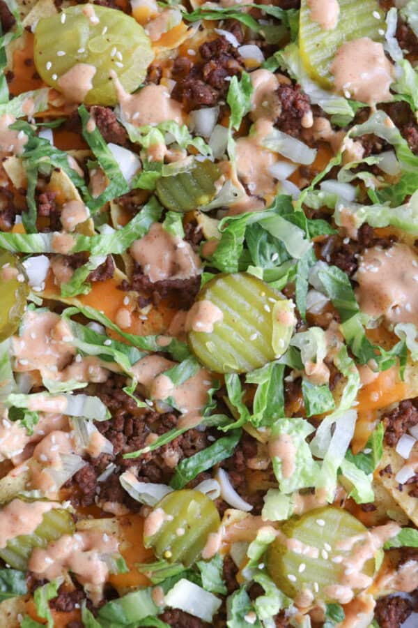 A close-up of nachos topped with ground beef, shredded lettuce, pickles, diced onions, melted cheese, sesame seeds, and a drizzle of creamy sauce.