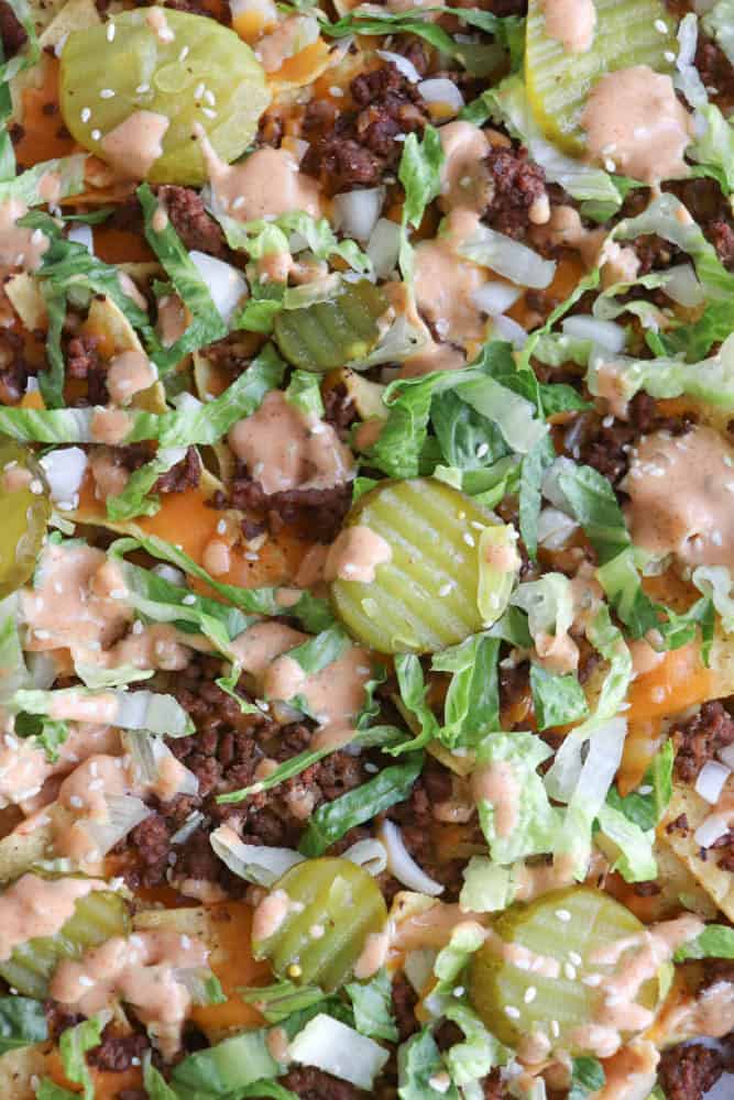 A close-up of nachos topped with ground beef, shredded lettuce, pickles, diced onions, melted cheese, sesame seeds, and a drizzle of creamy sauce.