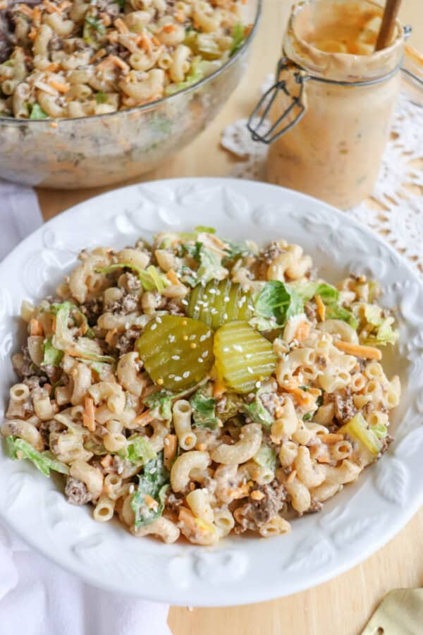 A white plate filled with macaroni salad mixed with lettuce, ground beef, shredded cheese, and topped with pickle slices. A glass bowl of more salad and a jar of creamy dressing are in the background.