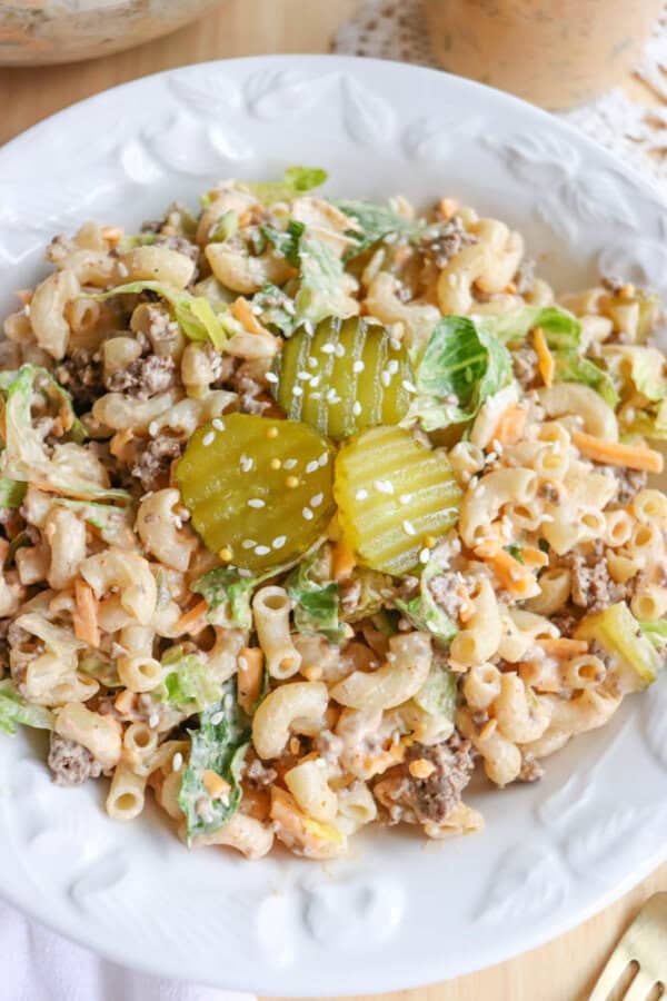 A white plate filled with macaroni salad mixed with ground beef, lettuce, shredded cheese, pickles, and sesame seeds, topped with three pickle slices in the center.
