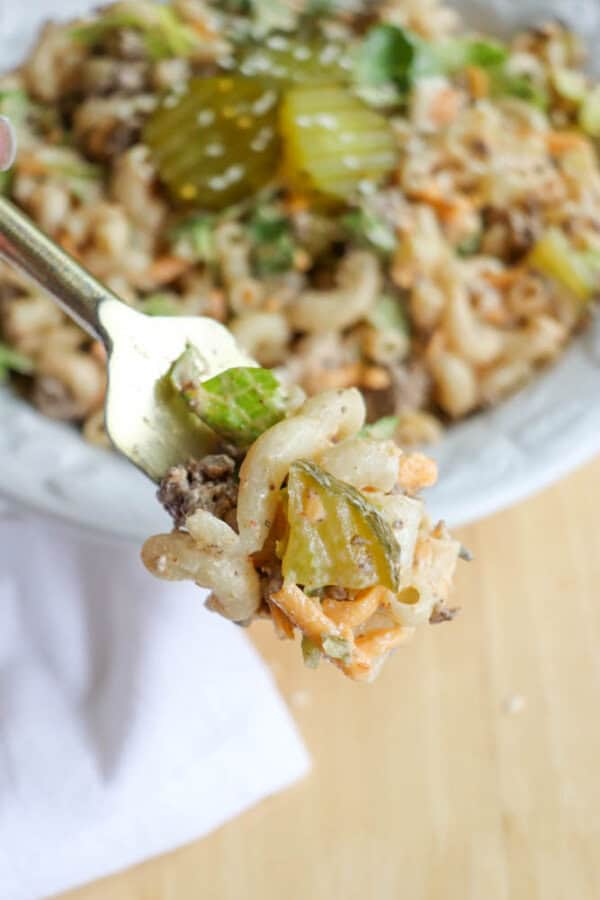 A close-up of a fork holding macaroni, ground beef, lettuce, shredded cheese, and a pickle slice, with a bowl of similar pasta salad in the background.