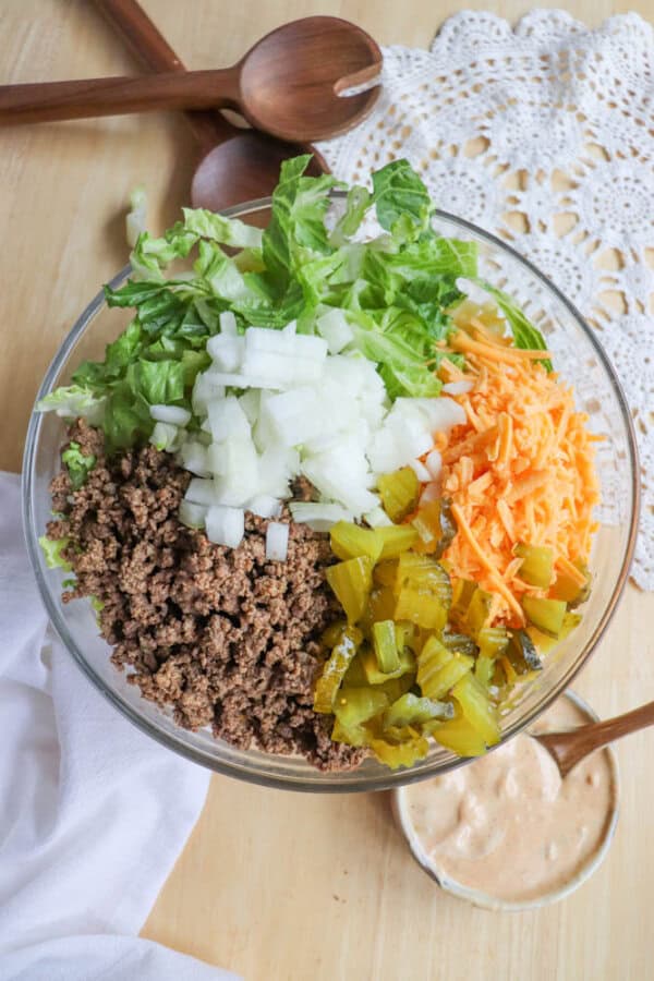 A glass bowl filled with chopped lettuce, diced onions, shredded cheddar cheese, cooked ground beef, and chopped pickles sits on a table, with a small bowl of sauce and wooden utensils nearby.