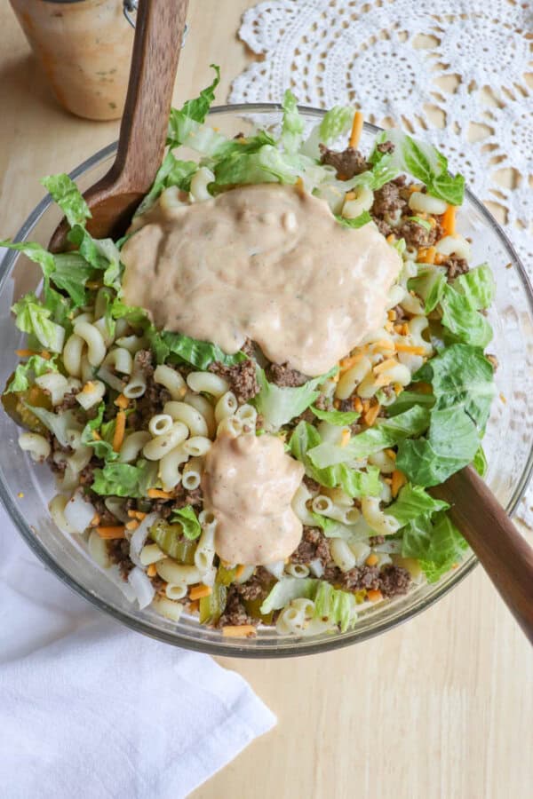 A glass bowl filled with a salad made of lettuce, macaroni, ground beef, shredded cheese, and topped with a creamy dressing, with wooden salad spoons resting inside the bowl.