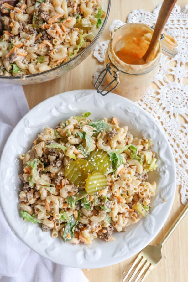 A white plate with macaroni salad topped with pickles. The salad includes lettuce, ground meat, shredded cheese, and sesame seeds. A fork is nearby, with a bowl of salad and a jar of creamy dressing in the background.