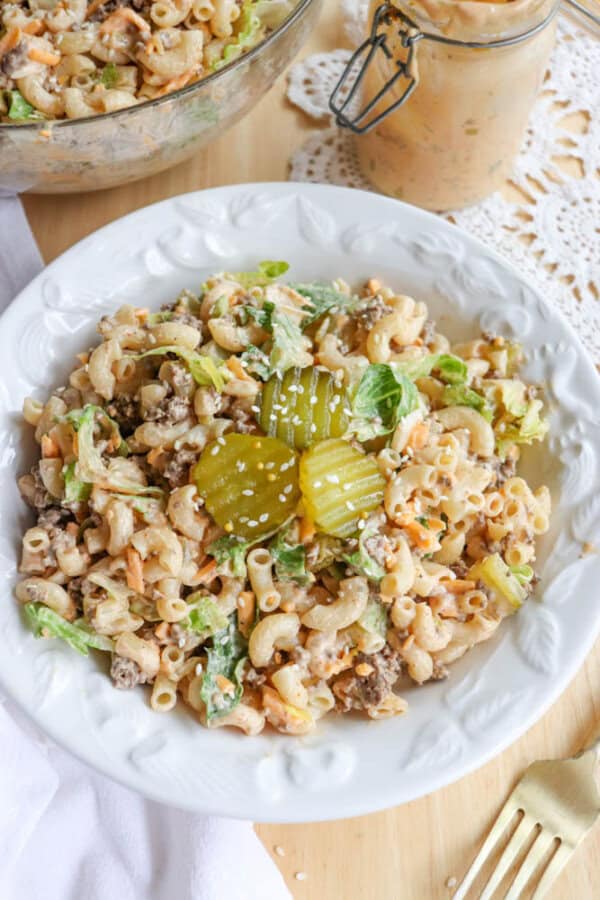 A white plate filled with macaroni salad topped with two pickle slices. The salad contains pasta, ground meat, lettuce, shredded carrots, and sesame seeds. A bowl and jar of extra salad are in the background on a wooden table.