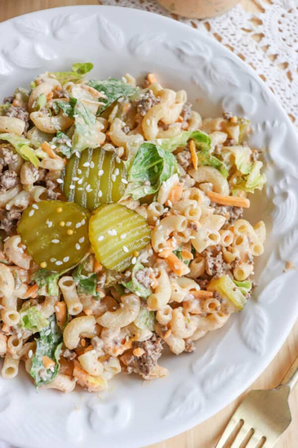 A white plate filled with macaroni salad mixed with ground beef, lettuce, and shredded cheese, topped with sliced pickles and sprinkled with sesame seeds. A gold fork and lace doily are nearby on the wooden table.