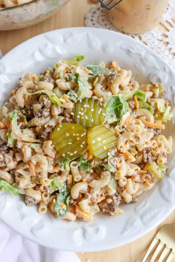 A white plate filled with macaroni salad mixed with ground beef, lettuce, shredded cheese, and dressing, topped with three pickle slices and sesame seeds. A gold fork is on the side.