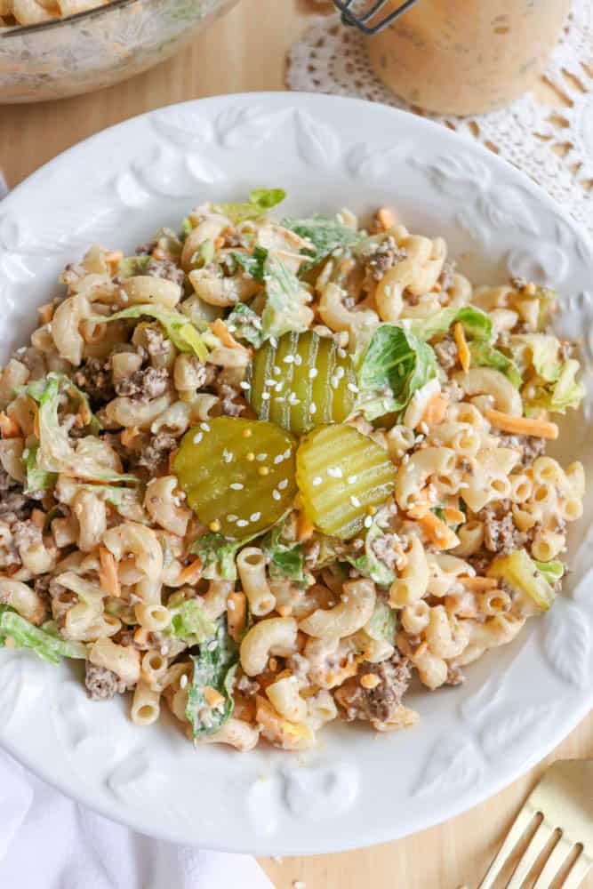 A white plate filled with macaroni salad mixed with ground beef, lettuce, shredded cheese, and dressing, topped with three pickle slices and sesame seeds. A gold fork is on the side.