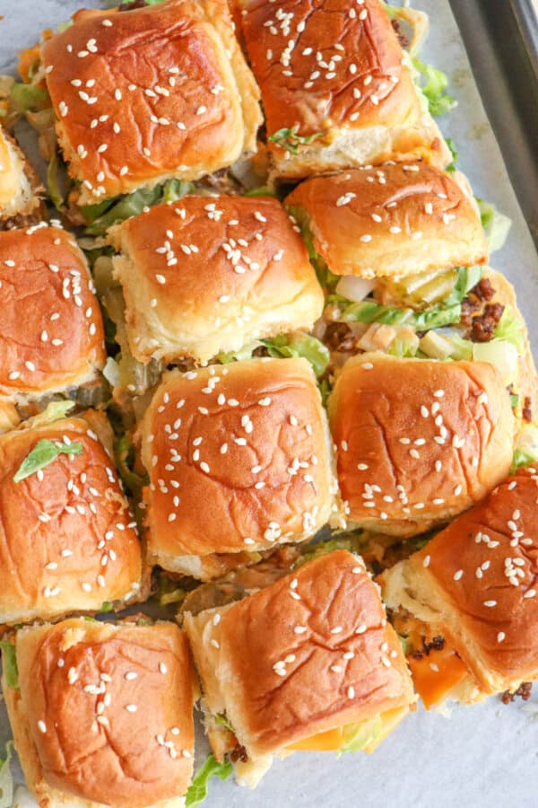 A tray of mini cheeseburger sliders topped with golden brown sesame seed buns, filled with lettuce, cheese, and ground beef, arranged closely together on parchment paper.