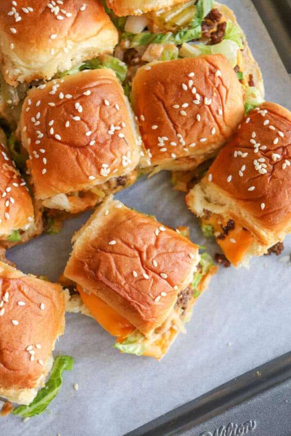 A close-up of several slider sandwiches with sesame seed buns, cheddar cheese, lettuce, and ground beef, arranged on a baking sheet lined with parchment paper.