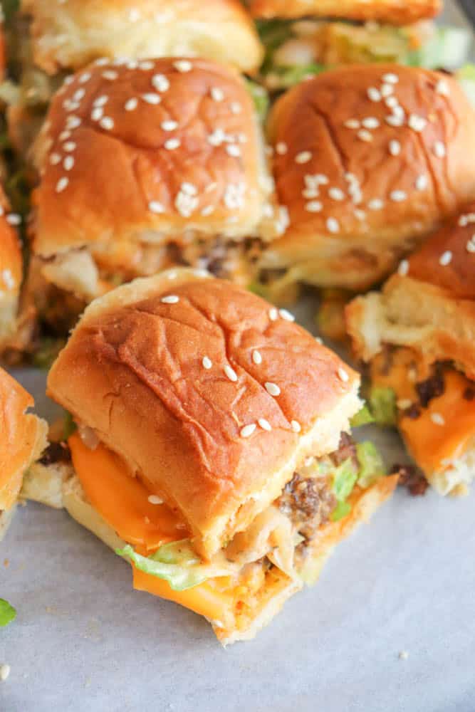 Close-up of several cheeseburger sliders on a tray, featuring golden-brown buns topped with sesame seeds, cheddar cheese, lettuce, and ground beef patties.