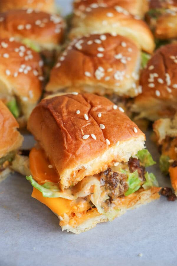 A close-up of mini cheeseburger sliders with lettuce, melted cheese, and ground beef filling, served on soft sesame seed buns. One slider is pulled slightly forward from the group.