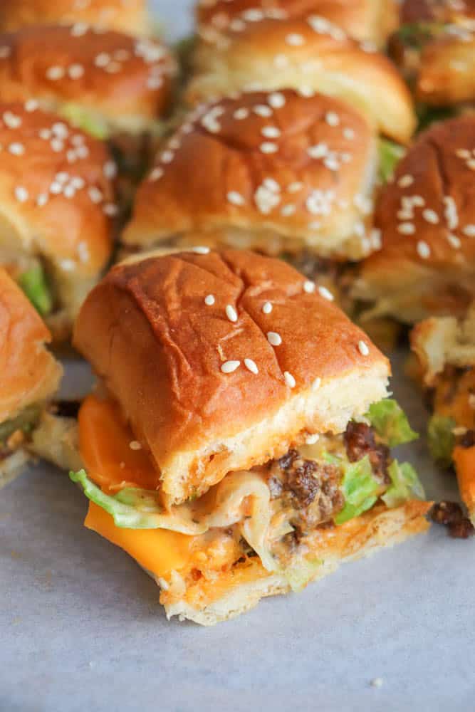 A close-up of mini cheeseburger sliders with lettuce, melted cheese, and ground beef filling, served on soft sesame seed buns. One slider is pulled slightly forward from the group.