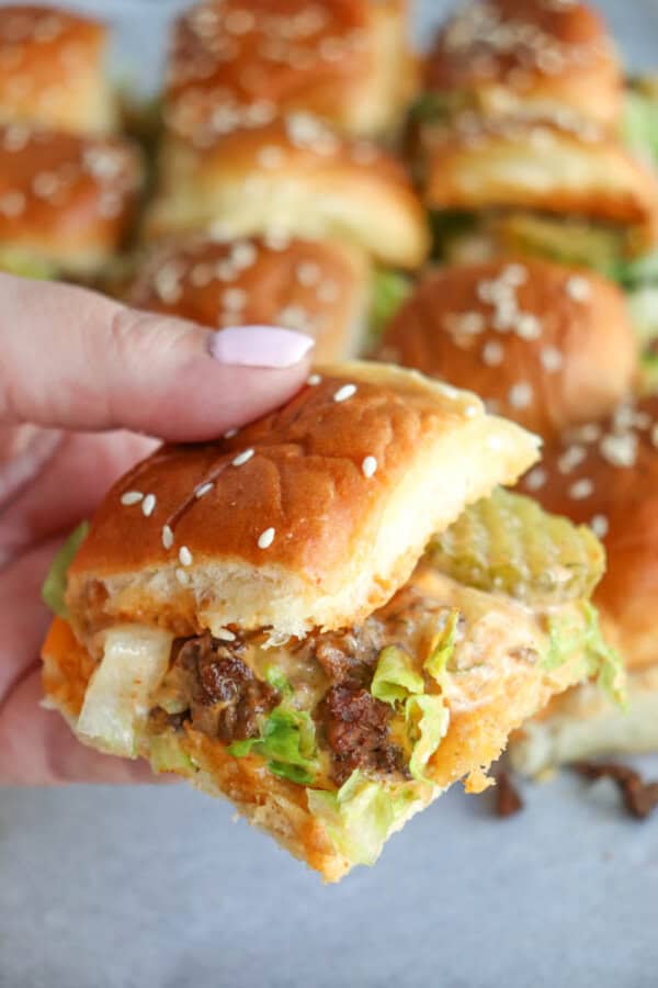 A hand holds a small cheeseburger slider with lettuce, melted cheese, pickles, and sauce in a sesame seed bun. More sliders are visible in the background.